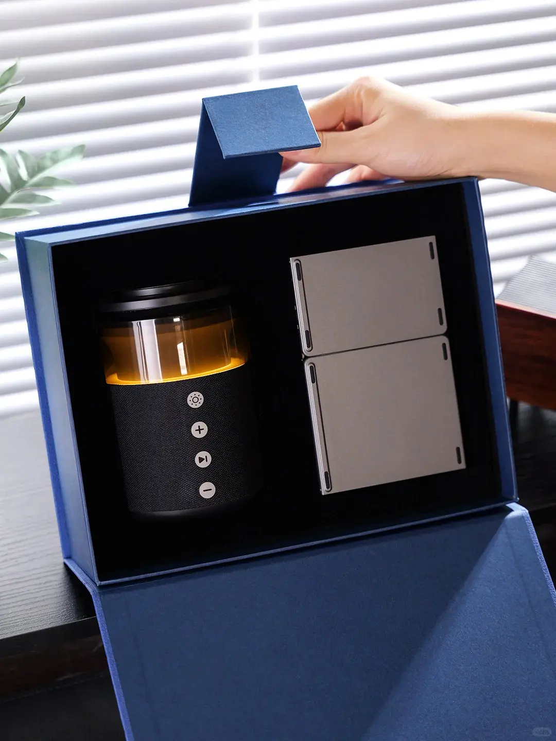 Person opening a tech-savvy corporate gift box with wireless charging speaker, 3-in-1 magnetic charger, and foldable wireless keyboard inside a navy blue box