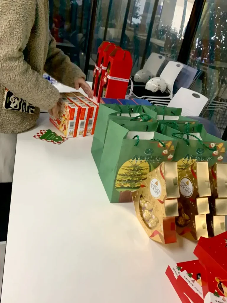 Office admin arranging 20 types of employee Christmas gifts including chocolates, mugs, Bluetooth speakers, snacks, and festive ornaments
