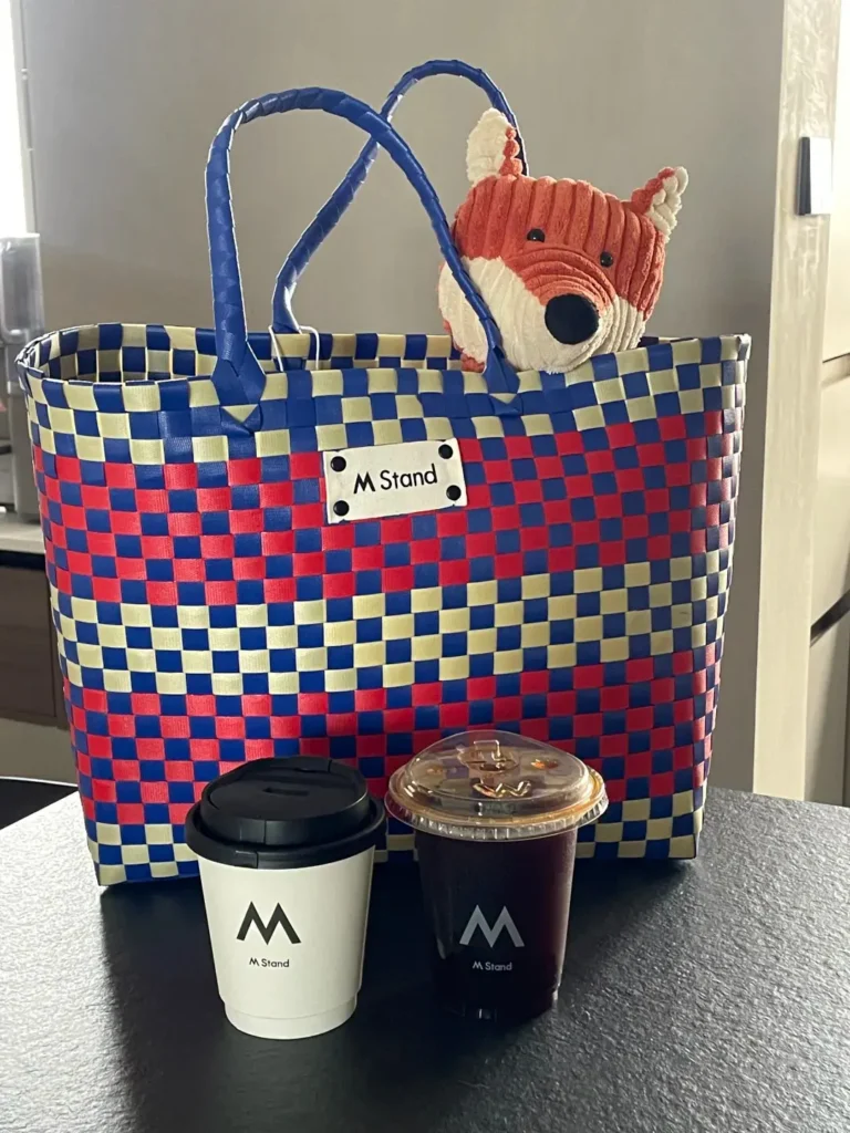 M Stand colorful plaid tote bag gift with purchase featuring a plush toy and two branded coffee cups displayed on a wooden table — a stylish and cozy GWP concept.