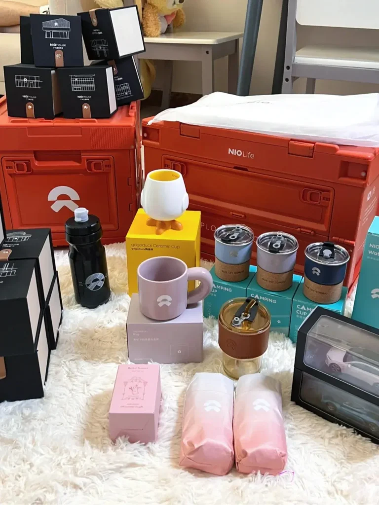 NIO gift with purchase collection displayed on a white blanket, featuring a camping storage box, foldable umbrella, ceramic mug, coffee cup, cosmetic pouch, toy car, and thermal bottle — stylish lifestyle gifts that enhance the brand experience.