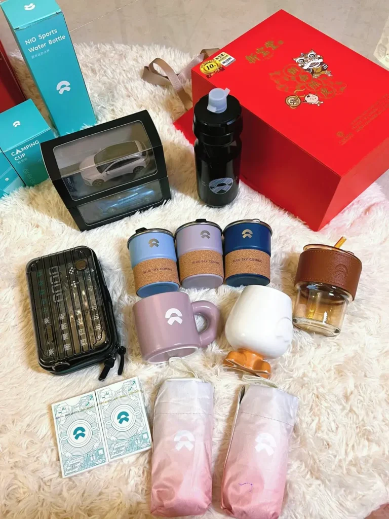 NIO gift with purchase ideas collection featuring branded umbrella, coffee mug, thermal bottle, cosmetic pouch, toy car, and playing cards — premium gifts that reflect the brand’s lifestyle values.