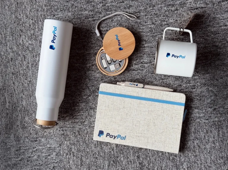 Company swag ideas for employees example featuring PayPal branded items including a tumbler, cable, pen, notebook and coffee mug